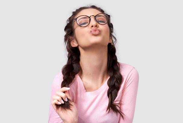 Closeup studio portrait of a pretty young European woman wearing pink long sleeve shirt, transparent eyewear, braids hairstyle, sending air kiss to his boyfriend while standing over white studio wall - Photo, Image