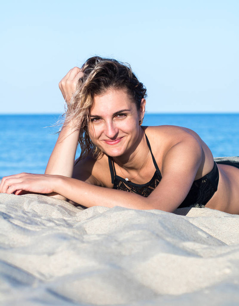 kumda siyah mayo kıvırcık saçlı tabaklanmış genç çekici kadın, kum üzerinde yalan - Fotoğraf, Görsel