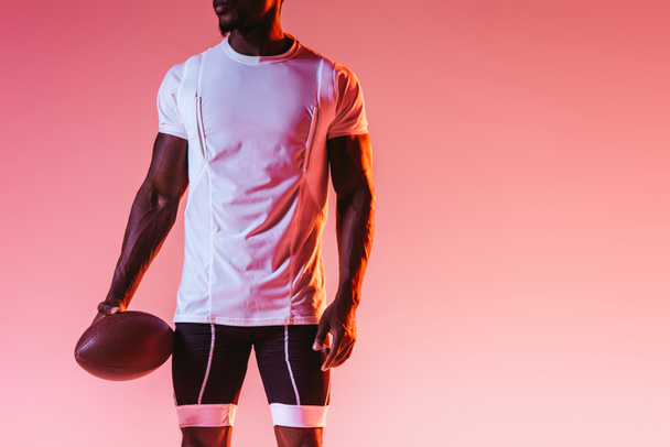 グラデーションと照明でピンクの背景にラグビーボールを保持するアフリカ系アメリカ人のスポーツマンのトリミングビュー - 写真・画像