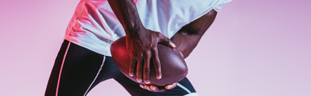 グラデーション、パノラマショットで紫色の背景にアメリカンフットボールをプレイアフリカ系アメリカ人のスポーツマンのトリミングビュー - 写真・画像