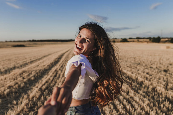 Follow me. Young woman holding hand and leading man to the beautiful nature sunset yellow landscape. View from back side, POV. Romantic couple travel, spend summer vacation together outdoors. - 写真・画像