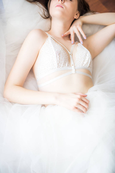 Young beautiful bride with dark long hair morning at home on a white bed in a white lace bustier and dress - Foto, Imagem