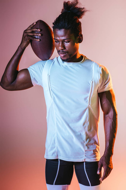 young african american sportsman holding rugby ball on pink background with gradient and lighting - Fotó, kép