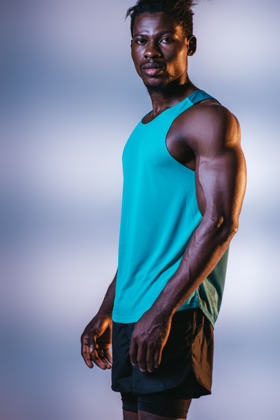 knappe, atletische Afro-Amerikaanse sportman kijkend naar camera op grijze en blauwe achtergrond met verlichting - Foto, afbeelding