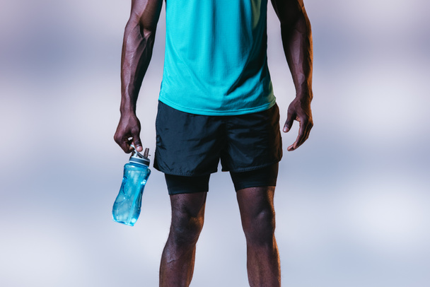 cropped view of african american sportsman in shorts holding sports bottle on grey background with lighting - Fotografie, Obrázek