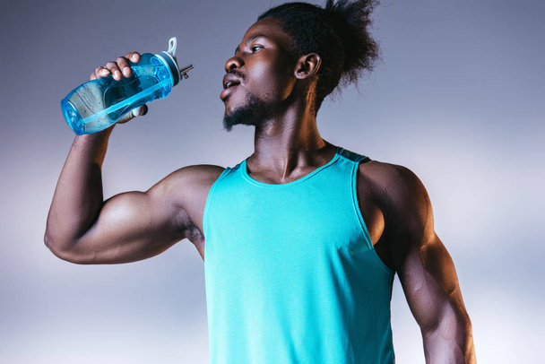 jonge, gespierde Afro-Amerikaanse sportman drinken uit sport fles op grijs en blauw gradiënt achtergrond met verlichting - Foto, afbeelding