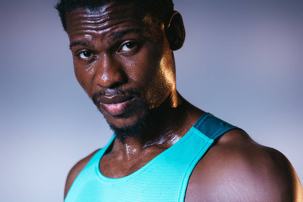 όμορφος, Αφροαμερικανός αθλητικός αθλητής με ιδρωμένη φάτσα κοιτάζοντας την κάμερα σε γκρι και μπλε φόντο ντεγκραντέ με φωτισμό - Φωτογραφία, εικόνα