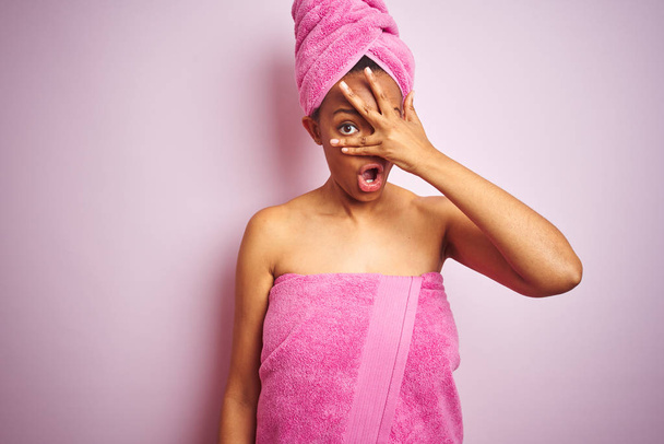 Afrikkalainen amerikkalainen nainen yllään suihku pyyhe kylvyn jälkeen yli vaaleanpunainen eristetty tausta kurkistaa shokki kattaa kasvot ja silmät käsin, katsellen sormien kautta nolostunut ilme
. - Valokuva, kuva