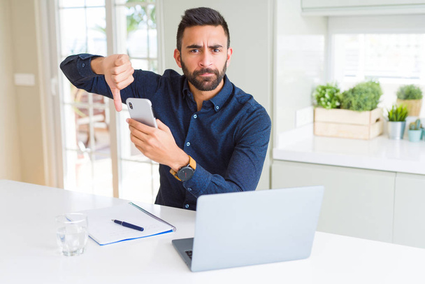 Handsome hispanic business man using smartphone and laptop at the office with angry face, negative sign showing dislike with thumbs down, rejection concept - Photo, Image