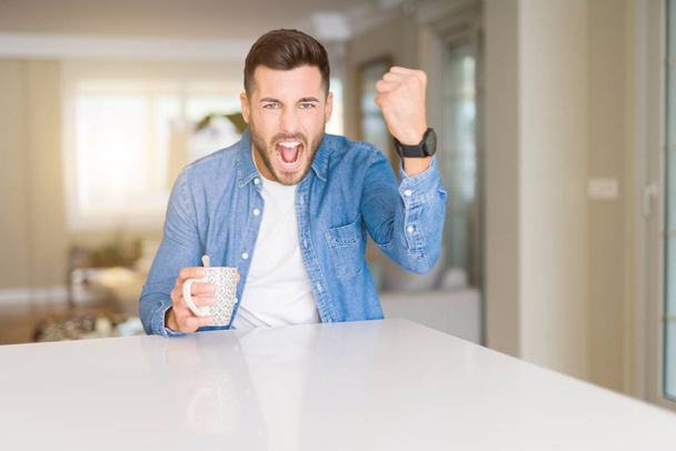 Giovane bell'uomo che beve una tazza di caffè a casa infastidito e frustrato gridando con rabbia, pazzo e urlando con mano alzata, concetto di rabbia
 - Foto, immagini