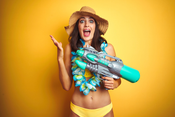 Hermosa mujer con bikini y flores hawaianas lei y pistola de agua sobre un fondo aislado muy feliz y emocionado, expresión ganadora celebrando la victoria gritando con una gran sonrisa y las manos levantadas
 - Foto, imagen