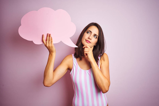 Beautiful woman wearing striped pink swimsuit and holding talking balloon over pink background serious face thinking about question, very confused idea - Photo, image
