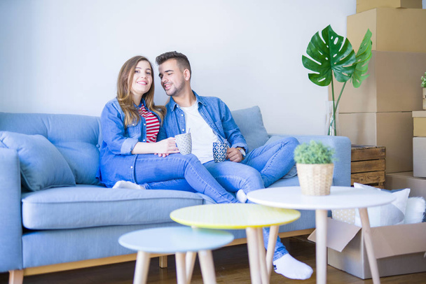 Young couple relaxing drinking a cup of coffee sitting at sofa of  new home with cardboard boxes around them, very happy moving to a new apartment - Foto, afbeelding