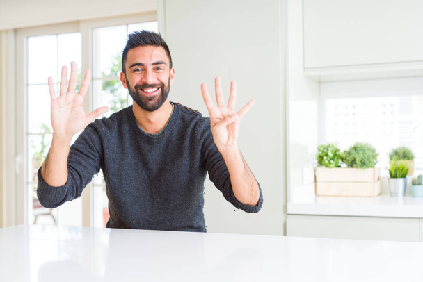 Knappe Spaanse man draagt casual trui thuis tonen en wijzend met de vingers nummer negen terwijl glimlachend zelfverzekerd en gelukkig. - Foto, afbeelding