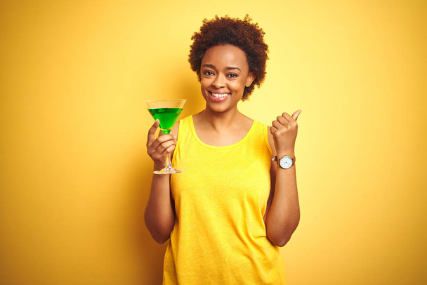 Jeune femme afro-américaine aux cheveux afro buvant un cocktail sur fond jaune isolé pointant et montrant le pouce sur le côté avec un sourire heureux
 - Photo, image