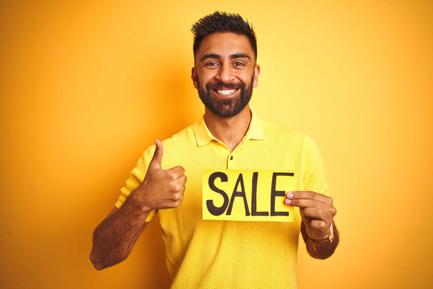 Jovem árabe indiano hispânico homem segurando banner venda sobre isolado fundo amarelo feliz com grande sorriso fazendo ok sinal, polegar com os dedos, excelente sinal
 - Foto, Imagem