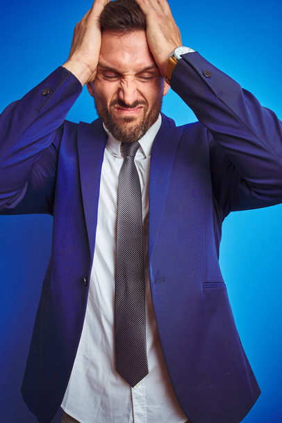 Imagen de ángulo vertical del joven hombre de negocios guapo sobre fondo azul aislado que sufre de dolor de cabeza desesperado y estresado debido al dolor y la migraña. Manos en la cabeza
. - Foto, imagen