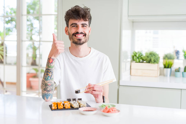 junger Mann isst asiatisches Sushi von zu Hause Lieferung glücklich mit großem Lächeln tun ok Zeichen, Daumen nach oben mit den Fingern, hervorragende Zeichen - Foto, Bild
