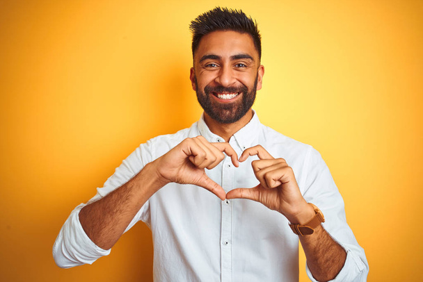 Ινδός επιχειρηματίας φορώντας λευκό κομψό πουκάμισο στέκεται πάνω από απομονωμένο κίτρινο φόντο χαμογελώντας στην αγάπη δείχνοντας το σύμβολο της καρδιάς και το σχήμα με τα χέρια. Ρομαντική ιδέα. - Φωτογραφία, εικόνα