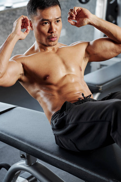 Handsome Vietnamese man doing abs workout on gym bench in fitness club - Zdjęcie, obraz