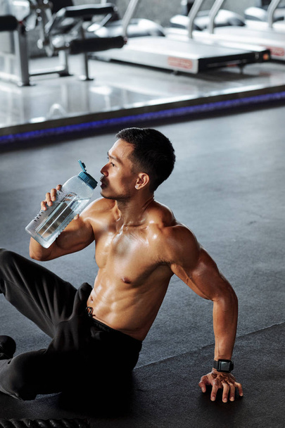 Мужчина без рубашки, сидящий на полу и пьющий воду из бутылки, отдыхающий после спортивных тренировок
 - Фото, изображение
