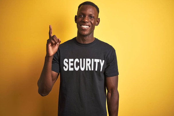 Afroamerický bezpečnostní muž, který nosí bezpečnostní uniformu nad izolovaným žlutým pozadím, ukázal prstem na úspěšnou myšlenku. Skončil a šťastný. Číslo jedna. - Fotografie, Obrázek