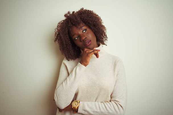 孤立した白い背景の上にタートルネックのセーターを着た若いアフリカのアフロ女性は、質問、ペンシブな表情を考えるあごに手を当てます。思慮深い顔で微笑む。疑いの概念. - 写真・画像