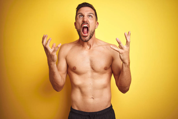 Young handsome shirtless man over isolated yellow background crazy and mad shouting and yelling with aggressive expression and arms raised. Frustration concept. - Foto, Bild