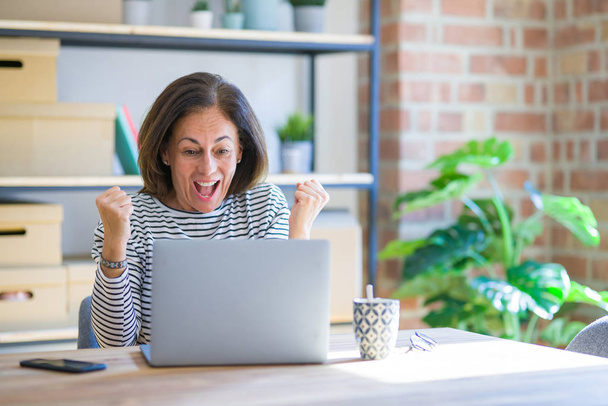 Femme âgée d'âge moyen assis à la table à la maison travaillant à l'aide d'un ordinateur portable criant fier et célébrant la victoire et le succès très excité, acclamant l'émotion
 - Photo, image
