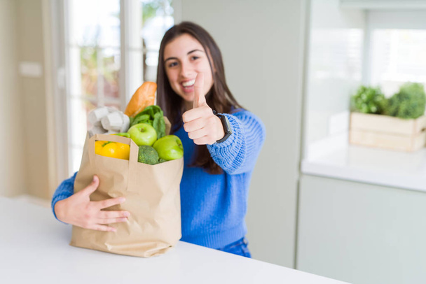 Красивая молодая женщина держит бумажный пакет, полный здоровых продуктов счастливой с большой улыбкой делает хорошо знак, палец вверх пальцами, отличный знак
 - Фото, изображение