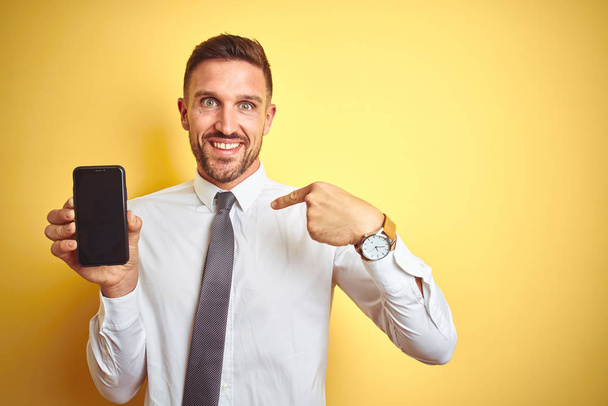 Молодой привлекательный бизнесмен показывает экран смартфона на желтом изолированном фоне с удивленным лицом, указывающим пальцем на себя
 - Фото, изображение