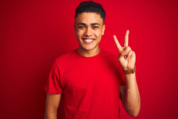 Jeune homme brésilien portant t-shirt debout sur fond rouge isolé montrant et pointant vers le haut avec les doigts numéro deux tout en souriant confiant et heureux
. - Photo, image