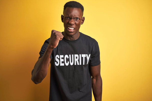 Afroamerický bezpečnostní muž, který nosí bezpečnostní uniformu nad izolovaným žlutým pozadím, rozzlobený a šílenou pěstí, frustrovaný a rozzuřený při křiku hněvem Zuřivost a agresivní koncepce. - Fotografie, Obrázek
