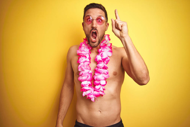 Young handsome shirtless man wearing sunglasses and pink hawaiian lei over yellow background pointing finger up with successful idea. Exited and happy. Number one. - Zdjęcie, obraz