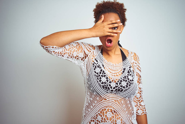 Молодая африканская американка с афроволосами в бикини на белом изолированном фоне, смотрящая рукой через пальцы с смущенным выражением лица и глаз.
. - Фото, изображение