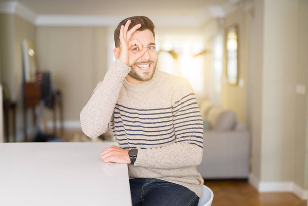 Joven hombre guapo usando un suéter en casa haciendo buen gesto con la mano sonriente, ojos mirando a través de los dedos con la cara feliz
. - Foto, imagen