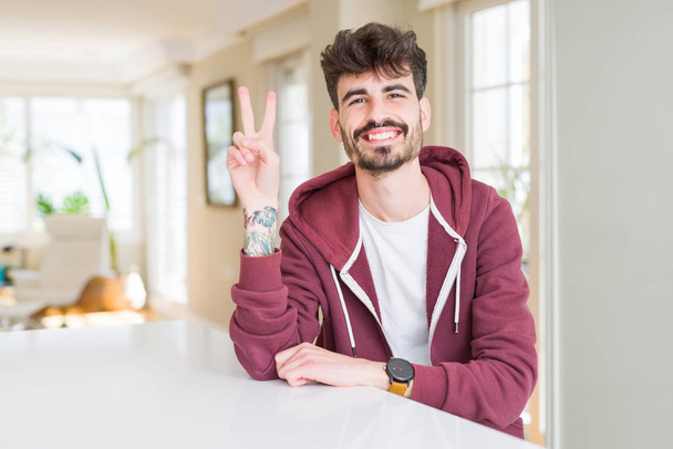 Young man wearing casual sweatshirt sitting on white table smiling with happy face winking at the camera doing victory sign. Number two. - Foto, imagen