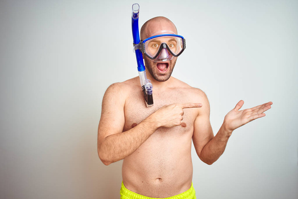 Jonge man het dragen van duik snorkel bril equipent over geïsoleerde achtergrond verbaasd en glimlachend naar de camera terwijl presenteren met de hand en wijzend met de vinger. - Foto, afbeelding