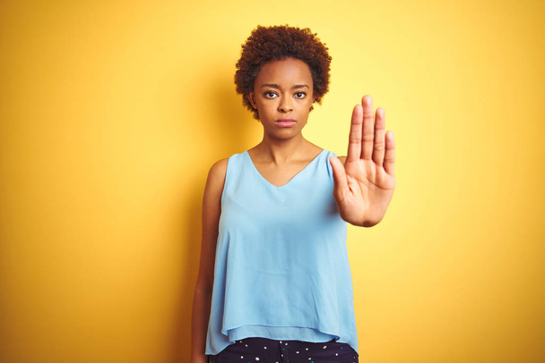 Красивая африканская американка в элегантной рубашке на изолированном жёлтом фоне делает остановку петь с ладонью руки. Предупреждающее выражение с негативным и серьезным жестом на лице
. - Фото, изображение