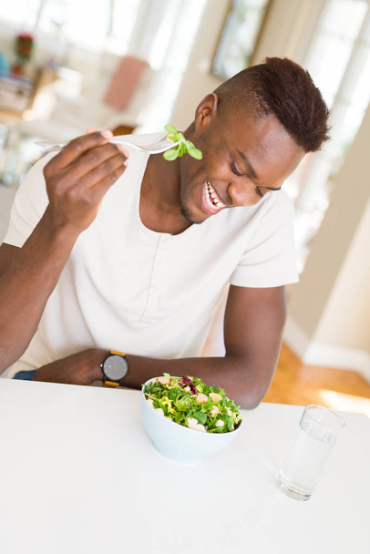 Marul, mutlu ve gülümseyerek masaya oturup yemek için çatal kullanarak sağlıklı sebze salata yemek Afrika yakışıklı genç - Fotoğraf, Görsel