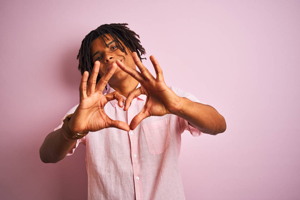 ドレッドロックを着たアフロアメリカンの男は、心のシンボルと形を手で示す愛に微笑む孤立したピンクの背景の上に立ってエレガントなシャツを着ています。ロマンチックなコンセプト. - 写真・画像