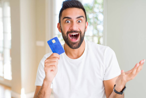 Hombre hispano guapo sosteniendo tarjeta de crédito muy feliz y emocionado, expresión ganadora celebrando la victoria gritando con una gran sonrisa y levantando las manos
 - Foto, Imagen