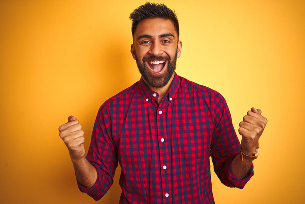 Молодой индиец в повседневной рубашке, стоящий на изолированном желтом фоне и радующийся удивлению и успеху с поднятыми руками и открытыми глазами. Концепция победителя
. - Фото, изображение