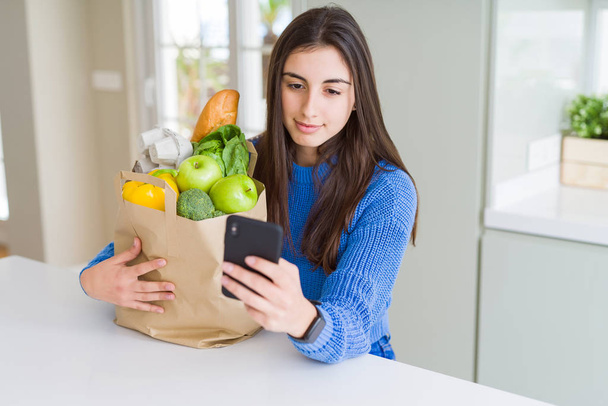 Young woman holding a paper bag full of fresh groceries and usin - Photo, Image