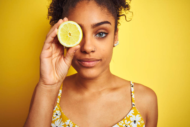 孤立した黄色の背景の上にレモンのスライスを保持する若いアフリカ系アメリカ人女性は、真剣に考えるスマートな顔に自信のある表情で - 写真・画像