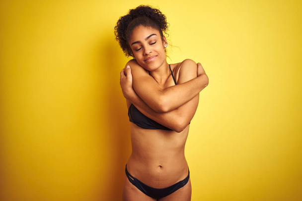 Afrikanische Amerikanerin im Urlaub im Bikini vor isoliertem gelben Hintergrund, die sich glücklich und positiv umarmt, selbstbewusst lächelnd. Selbstliebe und Selbstfürsorge - Foto, Bild