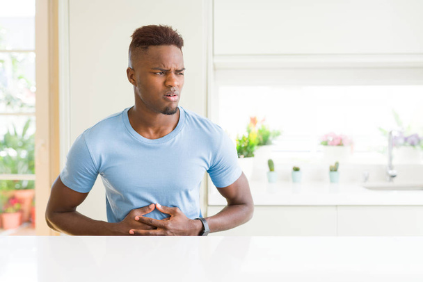 Όμορφος Αφρικανός Αμερικανός άνθρωπος φορώντας casual t-shirt στο σπίτι με το χέρι στο στομάχι, επειδή δυσπεψία, επώδυνη ασθένεια νιώθοντας αδιαθεσία. Αντίληψη του πόνο. - Φωτογραφία, εικόνα