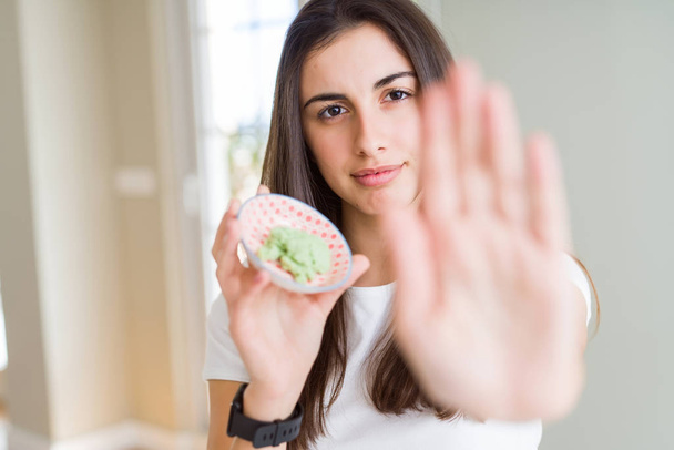 Beautiful young woman holding spicy asian wasabi with open hand doing stop sign with serious and confident expression, defense gesture - Foto, Imagem