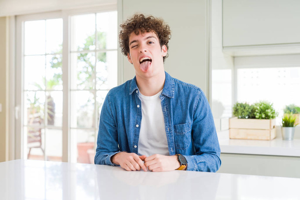 Young handsome man wearing casual denim jacket at home sticking tongue out happy with funny expression. Emotion concept. - Foto, Imagen