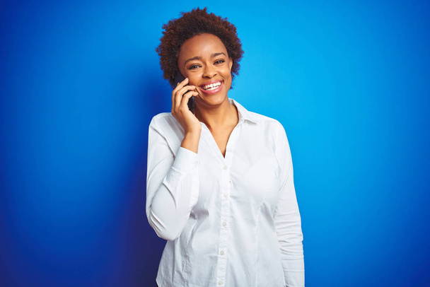 African american woman talking on the smartphone over blue isolated background with a happy face standing and smiling with a confident smile showing teeth - Fotografie, Obrázek